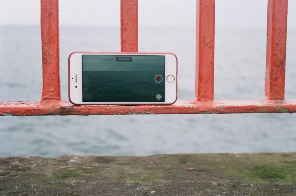 35mm photograph of a white iphone balanced on a red metal fence above a stone ledge, filming the sea and horizon line, which can be seen on the phones screen and out of focus behind the scene.