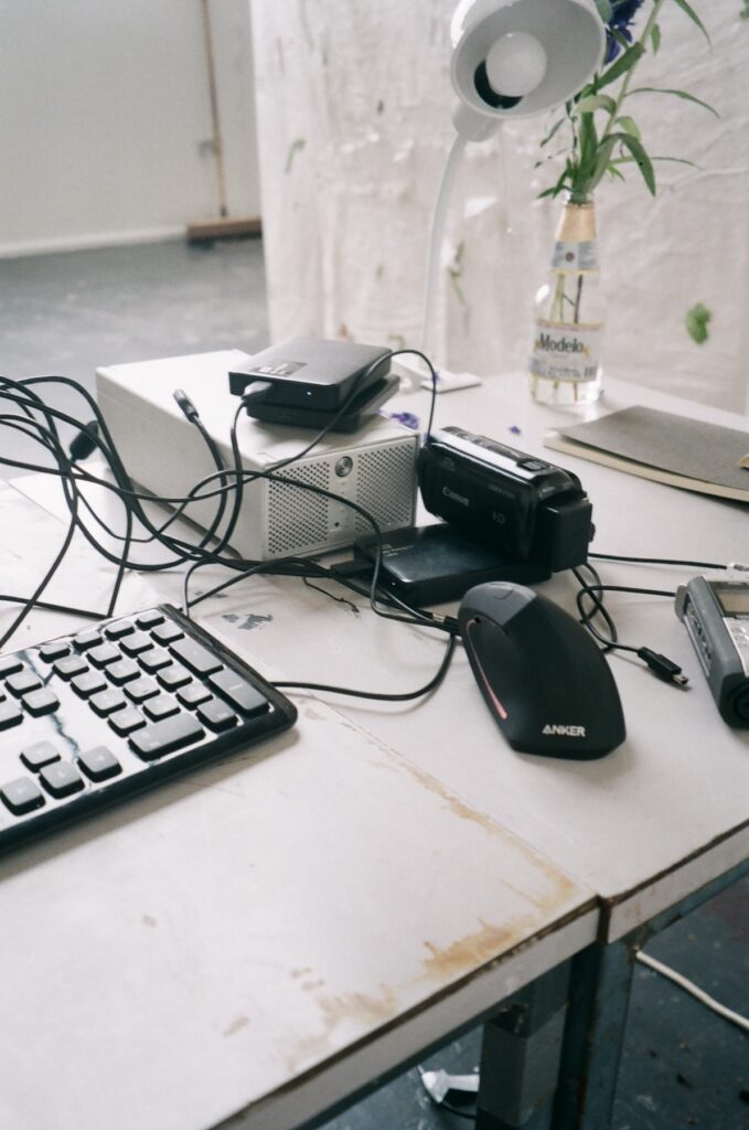 A white table holds a black keyboard, Anker wireless mouse, stacked electronic devices—including a hard drive and camcorder—and tangled black cables. A small white lamp and a clear vase with light purple flowers sit nearby. The table surface shows weathered off-white patches.