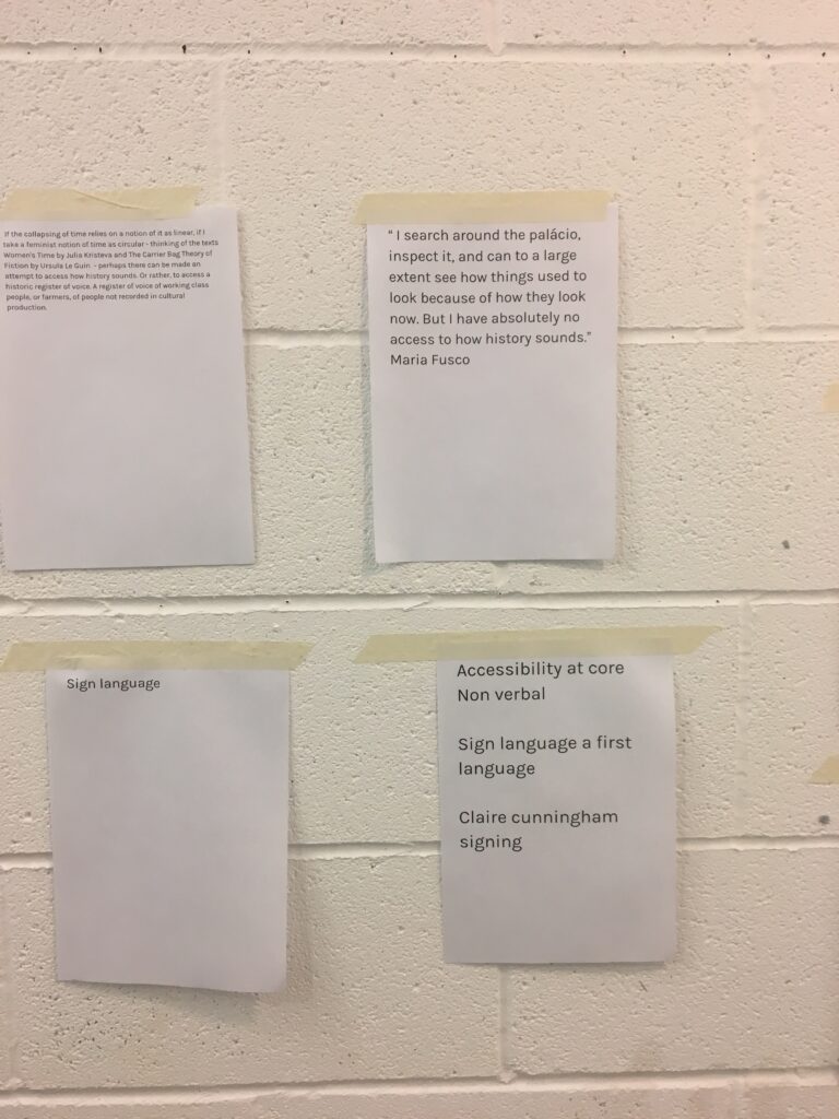 Four pieces of white paper with black text are taped to a light beige brick wall; they vary in size and content, including a paragraph beginning “I search around the palacio...” attributed to Maria Fusco, a sheet titled “Sign language”, and a list with phrases like “Accessibility at core Non Verbal”.