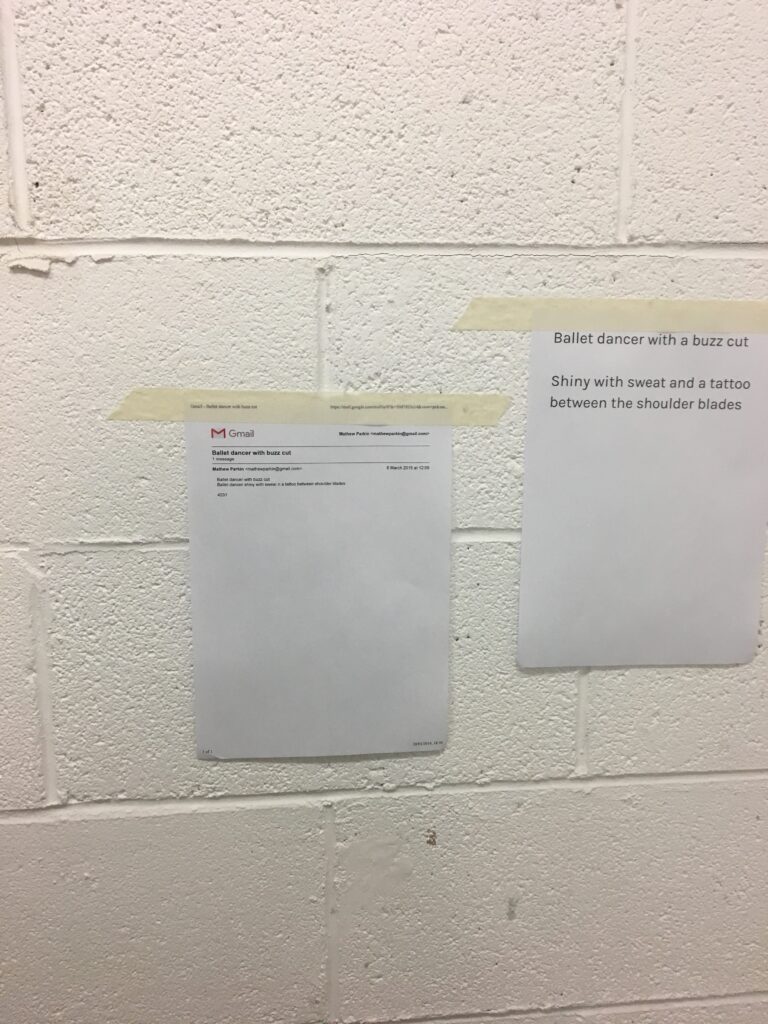 Two pieces of white paper are taped to a light beige cinder block wall with cream masking tape; one reads, “Ballet dancer with a buzz cut / Shiny with sweat and a tattoo between the shoulder blades”.