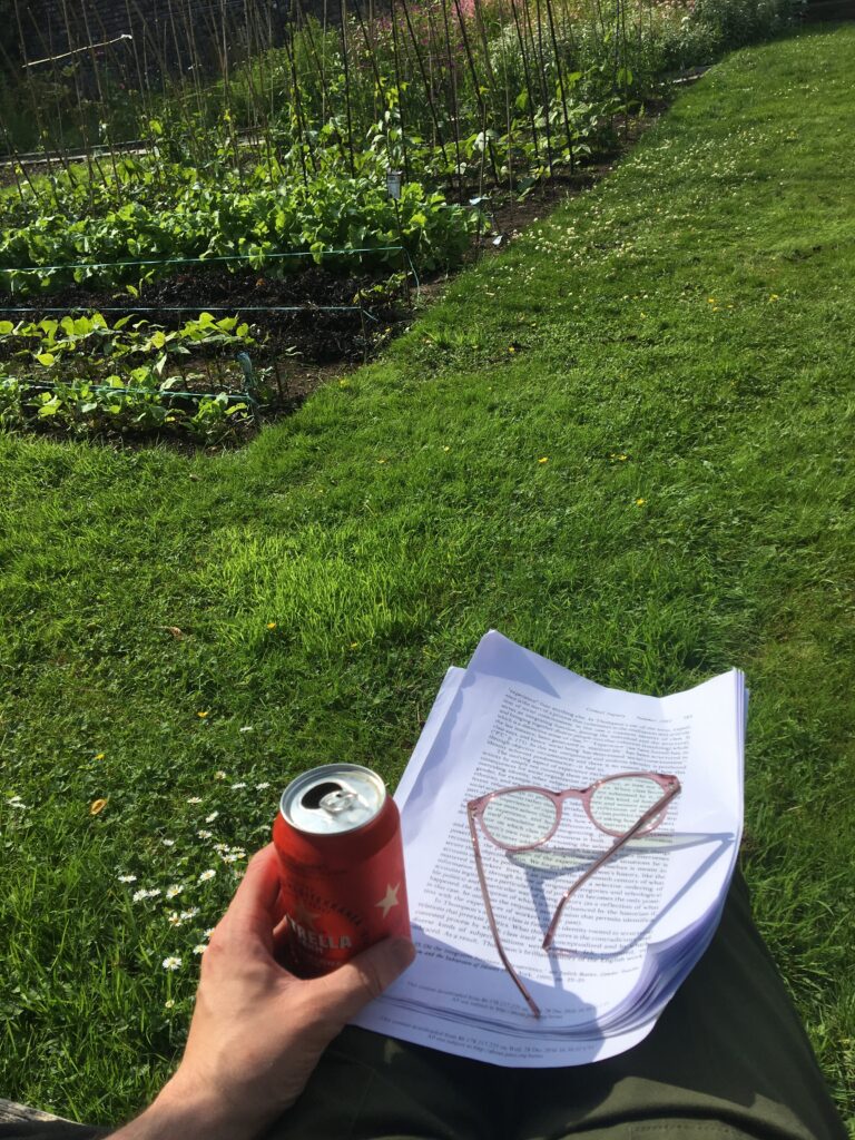 A person sits on grass holding a red can. Beside them, a stack of text-filled papers topped with pink round glasses rests on the ground. In the background, a garden with green plants, trellises, and white wildflowers is visible.
