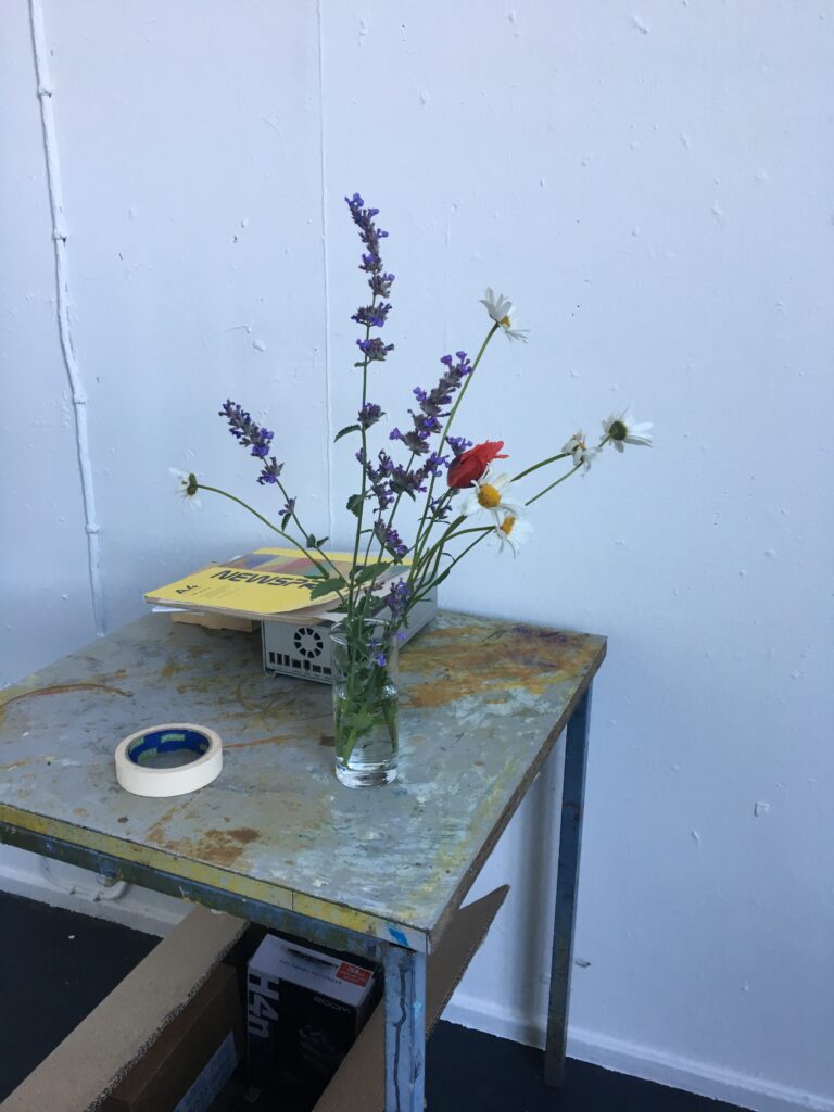 A clear glass vase on a weathered metal table holds wildflowers—lavender, white daisies, and a red poppy. A roll of masking tape and some printed paper peek out nearby. Cardboard boxes sit beneath the table against pale walls and a dark floor.