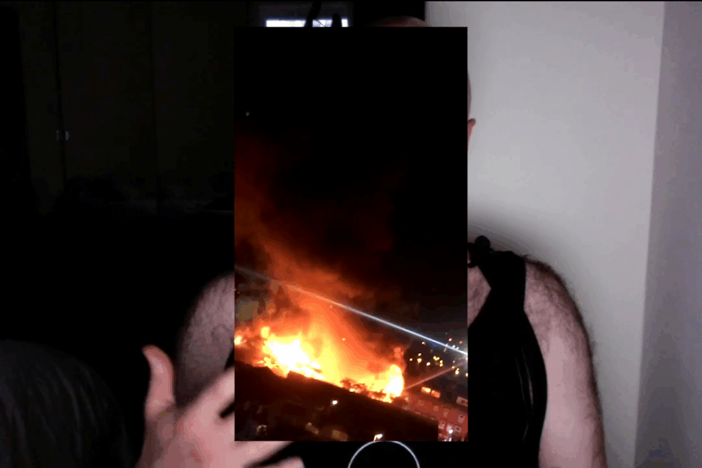 A webcam shot of light skinned hairy figure wearing a black vest is on webcam. Their face and part of their body is obscured by a portrait phone image overlaid of a vivid fire at nighttime.