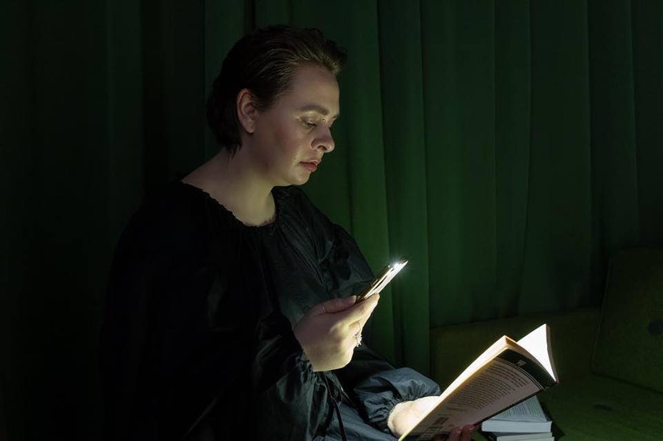 Charlotte Cullen in a black dress against a green curtain reading from a paperback book using a phone torch