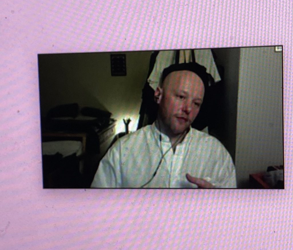 zoom window of myself with shaven head in white shirt talking against a pink background with pixels visible as a photo of a screen