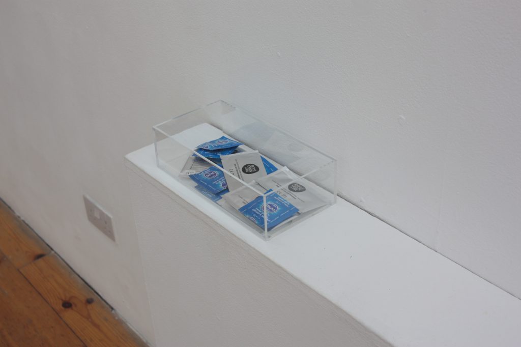 Perspex box on a white surface with sachets of condoms and lube