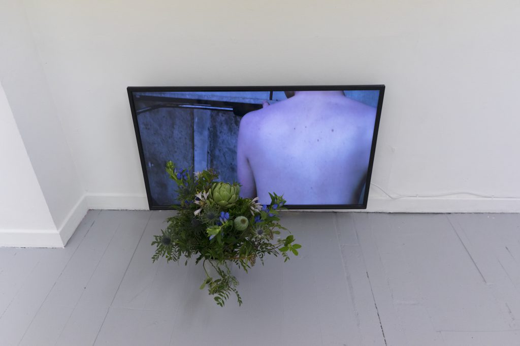 Screen sat on the floor with a mans back visible on it and a bouquet of flowers in front of it