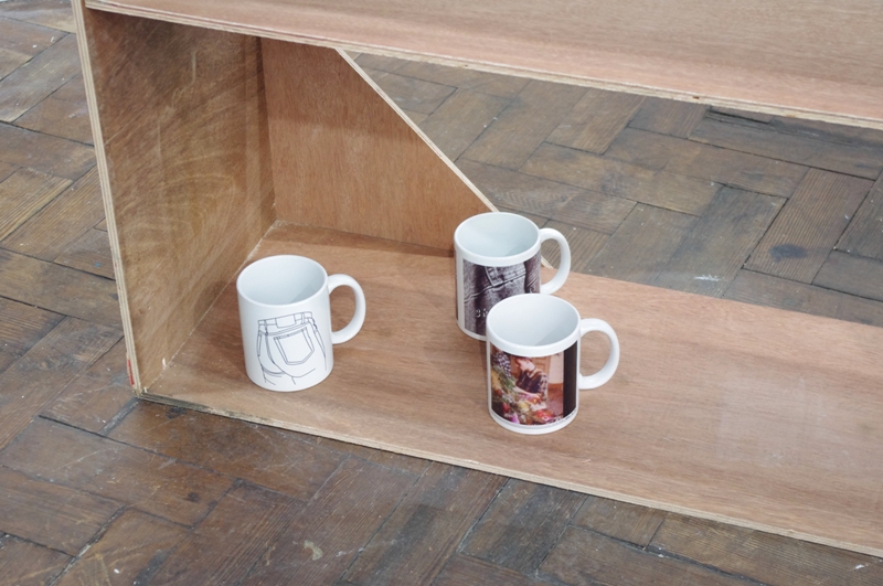 On a bottom plywood shelving structure on a parquet floor sit three white print on demand mugs. One has a line drawing of the ass of some jeans, one has a still from a 1970's Jean-Daniel Cadinot film, and a photograph of some denim wear.