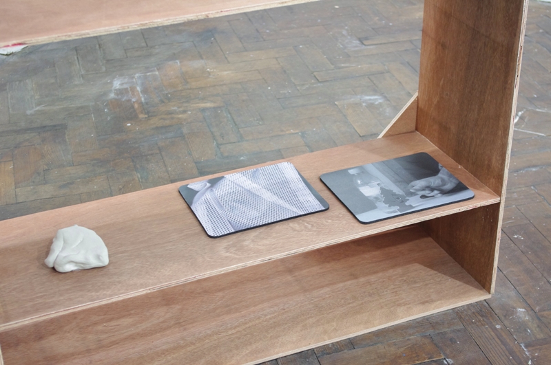 On a bottom plywood shelving structure on a parquet floor sit two mousemats and a lump of handled clay. One mousemat is the scanned lining of a sports jacket and the second is a black and white still life photograph.