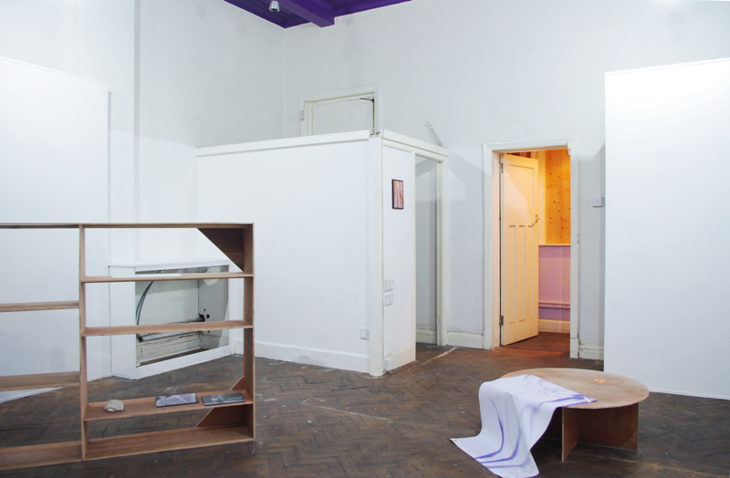 Empty looking white gallery with a parquet floor, door open and light on at the back. In the front is a freestanding plywood shelving structure with some mousemats on it. To its left is a squat circular platform with a textile hanging off it. 