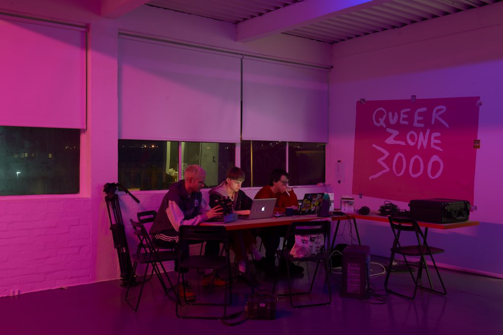 Pink image of three people sat at a large desk full of electronic equipment with a poster painted with QUEERZONE3000 behind them