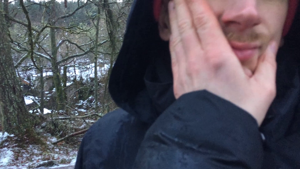 close up of a hand holding a mans hooded moustached face with snowy trees in the background