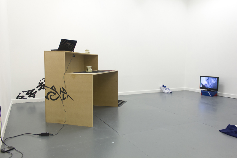 White gallery with a wooden desk with oil burners and a laptop on it. On the wall behind is a kappa logo, trainers, and a screen on a shoe box.