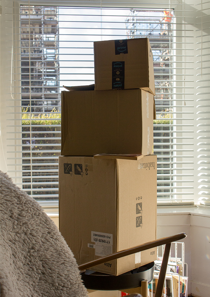 Cardboard box tower in front of a window with blinds with the edge of an armchair in the foreground 