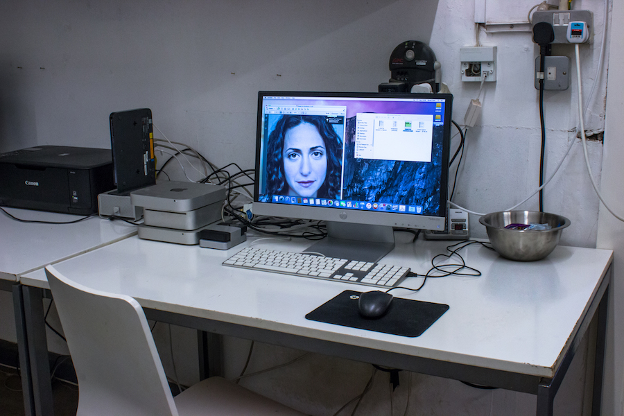 iMac on a white desk with various hard drives, Mac Mini's and a metal bowl of condom and lube