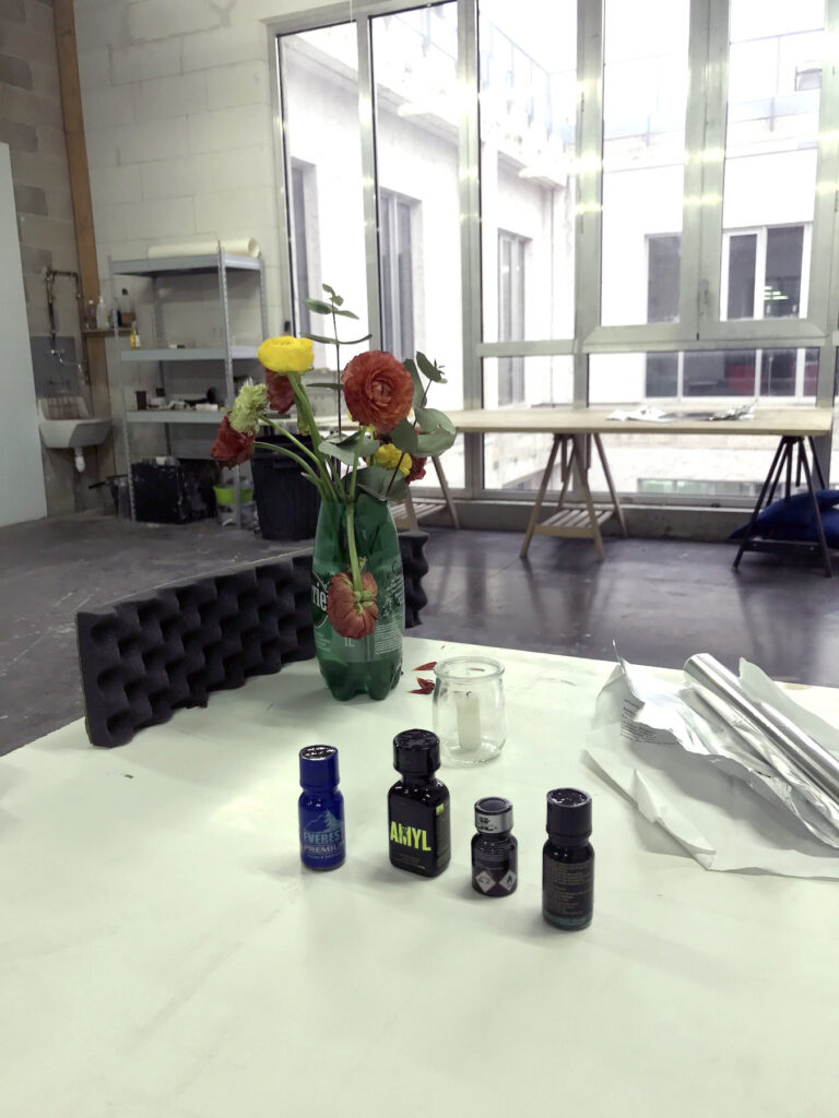 photo of studio desk with flowers in a plastic water bottle and a row of poppers