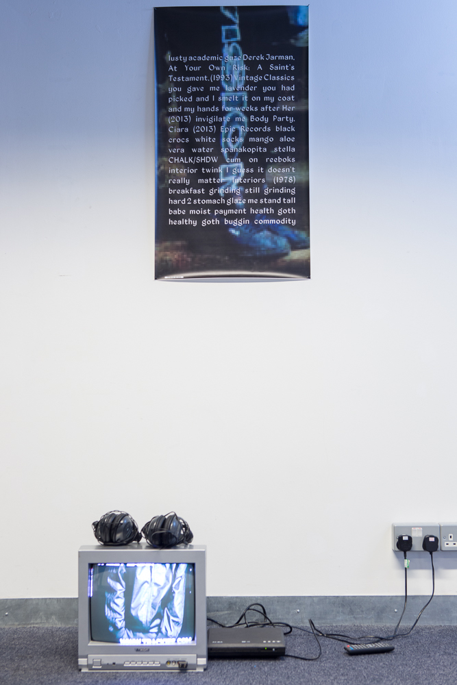 Banner on a white wall with a pixelated image of a leg in jogging bottoms overlaid with text, on the floor below the banner is a silver tv with headphones playing a video.