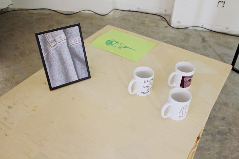 a framed photo of some blue denim, three white print on demand mugs, and a painting of a face on green paper on a wooden desk.