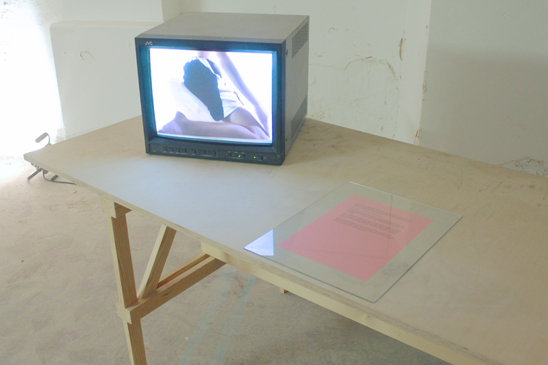 a cube monitor and a pink piece of paper with writing under a piece of glass, both on a wooden table.