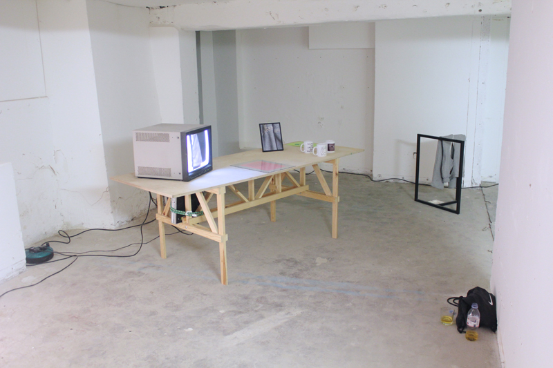 Wooden table with a tv, mugs, photos and papers on it in a white basement. A metal frame with grey joggers is in the back, and a black nike bag is to the right with a evian bottle with a yellow liquid. 