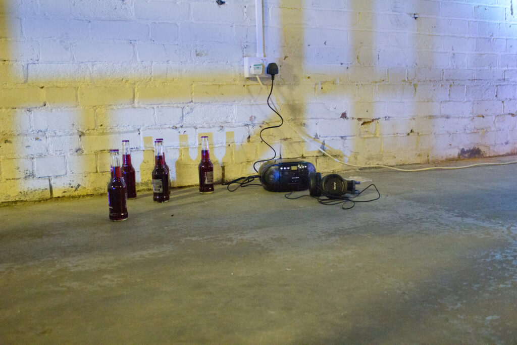 On a grey gallery floor against a white brick wall is a cheap black boombox with headphones plugged in surrounded by bottles of purple WKD alcopop.