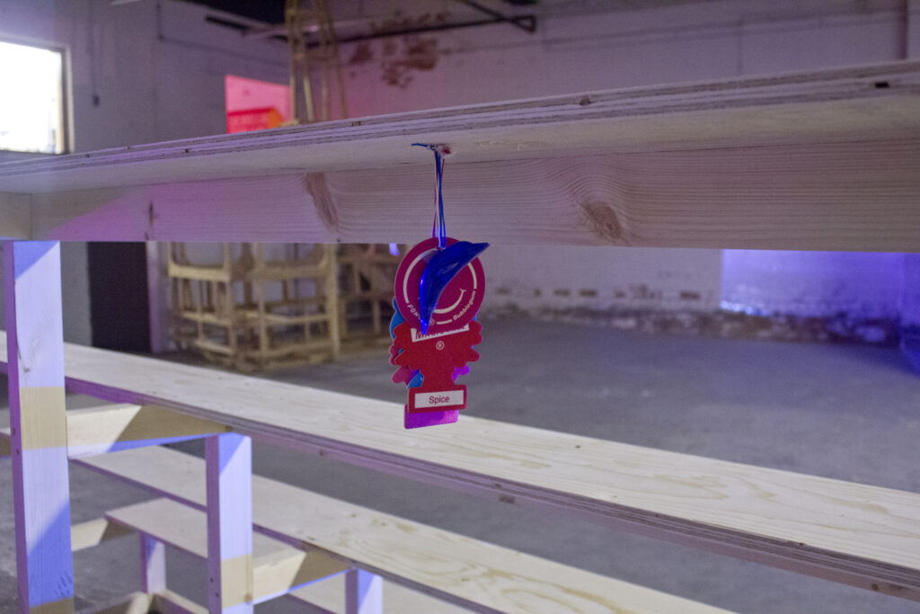 On the underneath or a plywood bleacher structure are tapped a bundle of several car air fresheners, visible is a blue dolphin, a pink smiley face and a red pine tree.