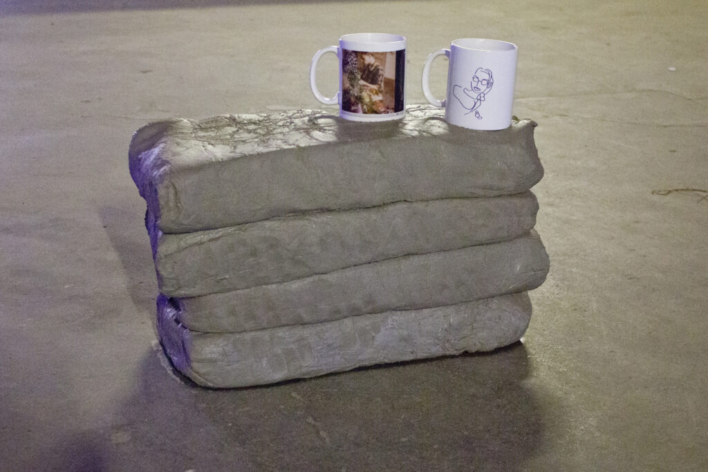 On a grey gallery floor four slabs of grey clay are piled on top of each other to create a plinth on top of which sits two white mugs one printed with a rough line drawing of Lara Croft, and the other with a vhs screenshot of twinks around a table doing flower arranging. 