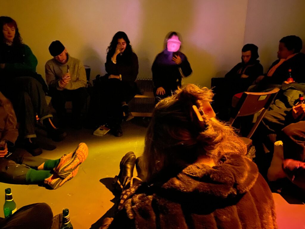 A group of people sit in a white gallery with low lighting making the space look orange, on a range of benches, chairs and the floor. There is a person reading on their phone which illuminates their face into a pink blur.