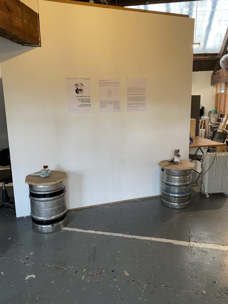 A white stud wall in a studio space with three white sheets of A3 with text on the pinned to the middle of the wall. Either side is wooden topped metal barrels with fabric and wire apple dolls on top.