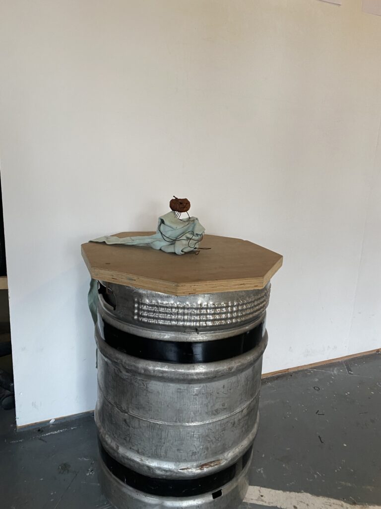 A wooden topped metal barrels with seafoam fabric and wire apple dolls on top in front of a white wall.