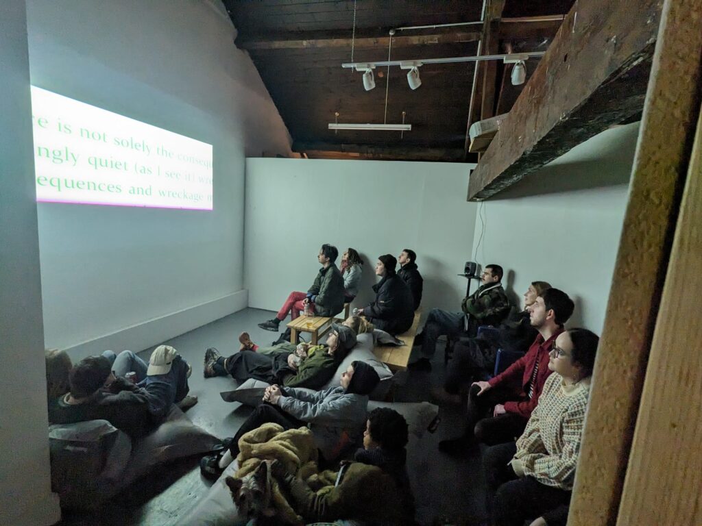 A group of people to the right of the image sit on benches and beanbags in a white gallery space looking at a video projected of a wall on the left of the image, due to the projection they are bathed in a blue light.