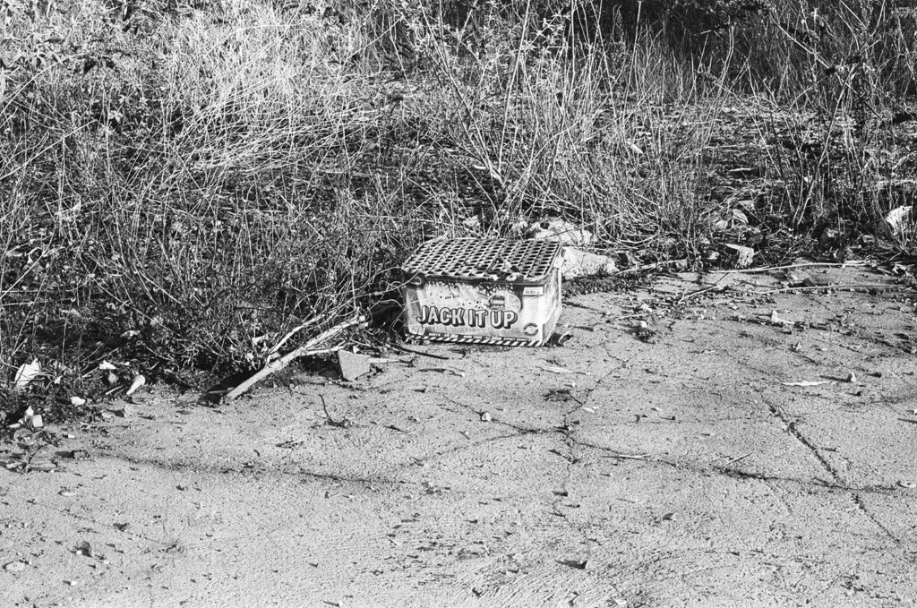 Black and white 35mm photograph of an overgrown wasteland with plastic firework container in the foreground with the text 'JACK IT UP'.