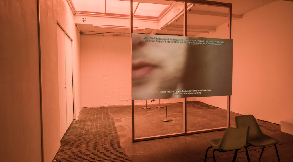 Installation image of a white gallery with orange lighting and a large screen splitting the space into two. In the foreground are green chairs tied together like loveseats. On the screen is a blurry mouth on a light skinned chin with a caption below reading ‘[‘Better off Alone’ by Alice Deejay plays softly in the background, sounds of a camera being handled]’, and at the top of the screen ‘[North West English Accent] A pair of lips and a light skin jaw cropped by the camera mouth the words to Alice Deejay’s ‘Better off Alone’ and drinking beer wearing a visible three stripes adidas top’.