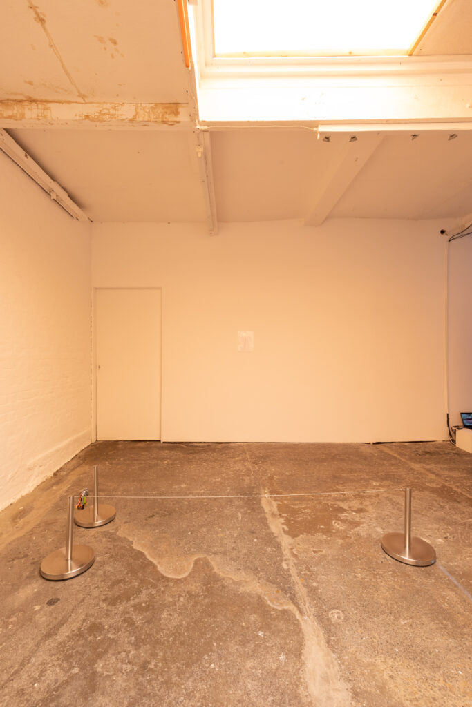 Installation image of a white gallery with orange lighting. In the foreground is a low uneven sculpture made from metal gallery barriers and an assortment of keyring and padlocks. On the back wall is paper in a plastic wallet pinned up. The floor is a rough grey concrete.