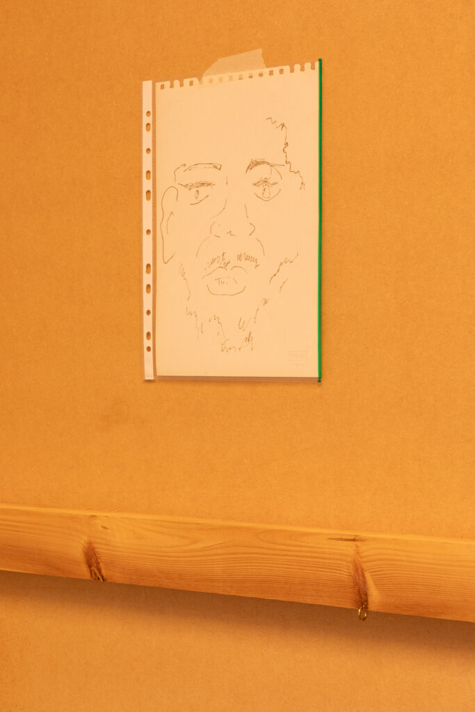 On a wooden surface is an a4 expressionistic drawing in gold pen of a mans face attached to the wall in a plastic wallet with a strip of masking tape. Below left a horizontal beam of wood has a gold earring piercing it where a nipple would be.