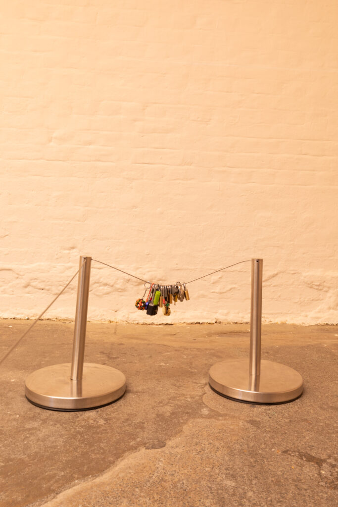 Installation image of a detail shot of a sculpture in a white gallery with orange lighting and a rough grey concrete floor. Between two metal gallery barriers the wire is sagging during to the weight of an assortment of keyring and padlocks bunched together.