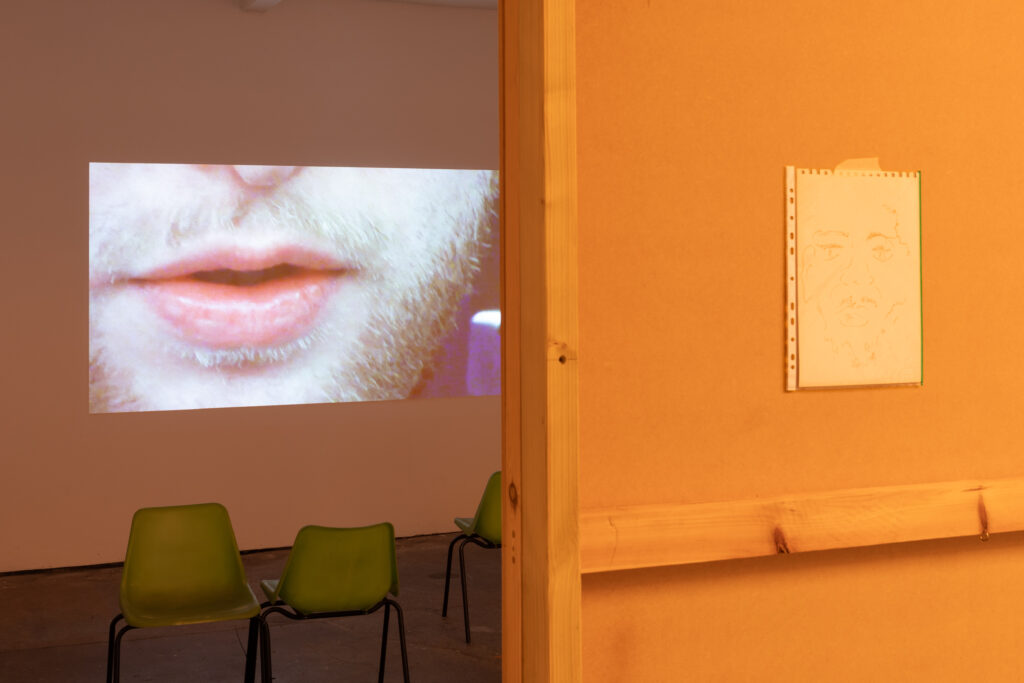 Installation image of a white gallery with orange lighting and a large wooden screen splitting the space into two. We took from the back of the screen to a projection on a wall of a pair of lips and a light skin jaw cropped by the camera in front of green chairs tied together like loveseats. On the wooden wall s an a4 expressionistic drawing in gold pen of a mans face attached to the wall in a plastic wallet with a strip of masking tape. Below left a horizontal beam of wood has a gold earring piercing it where a nipple would be.