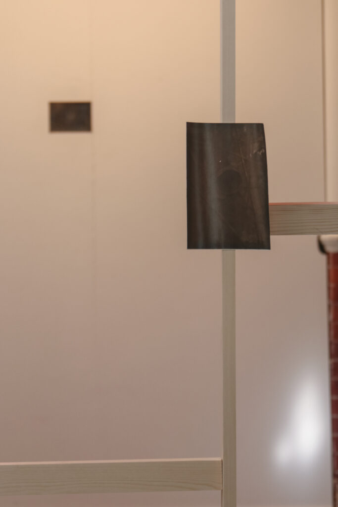 In a white gallery space, a dark hand printed photo of a pipe is attached to a light wooden frame.