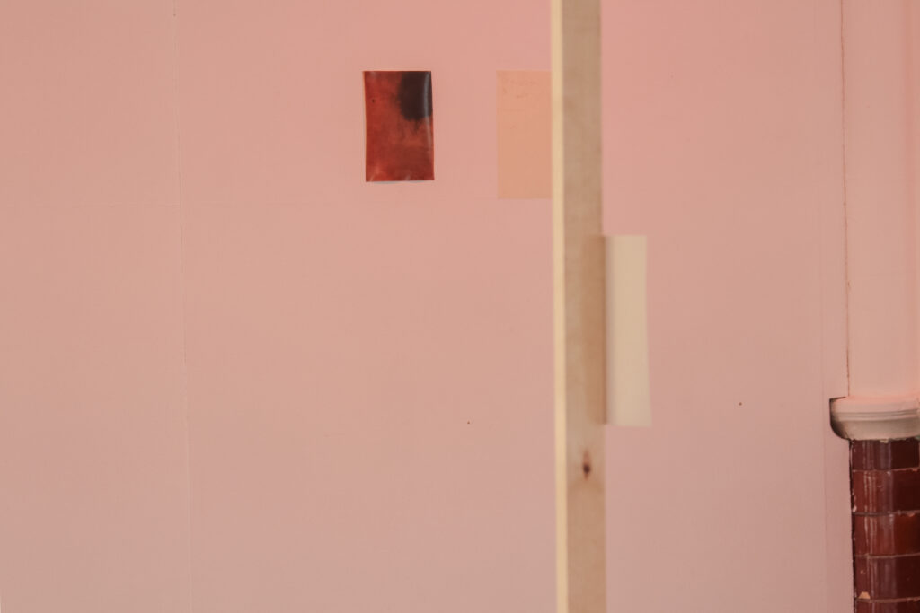 Installation shot of a thin wooden fame with hand printed photographs in a range of clarity due to differing development and paper types attached to the frame in a white gallery with subtle red lighting and some patches of red brick wall. On the back wall hangs a photograph next to a drawing on paper.