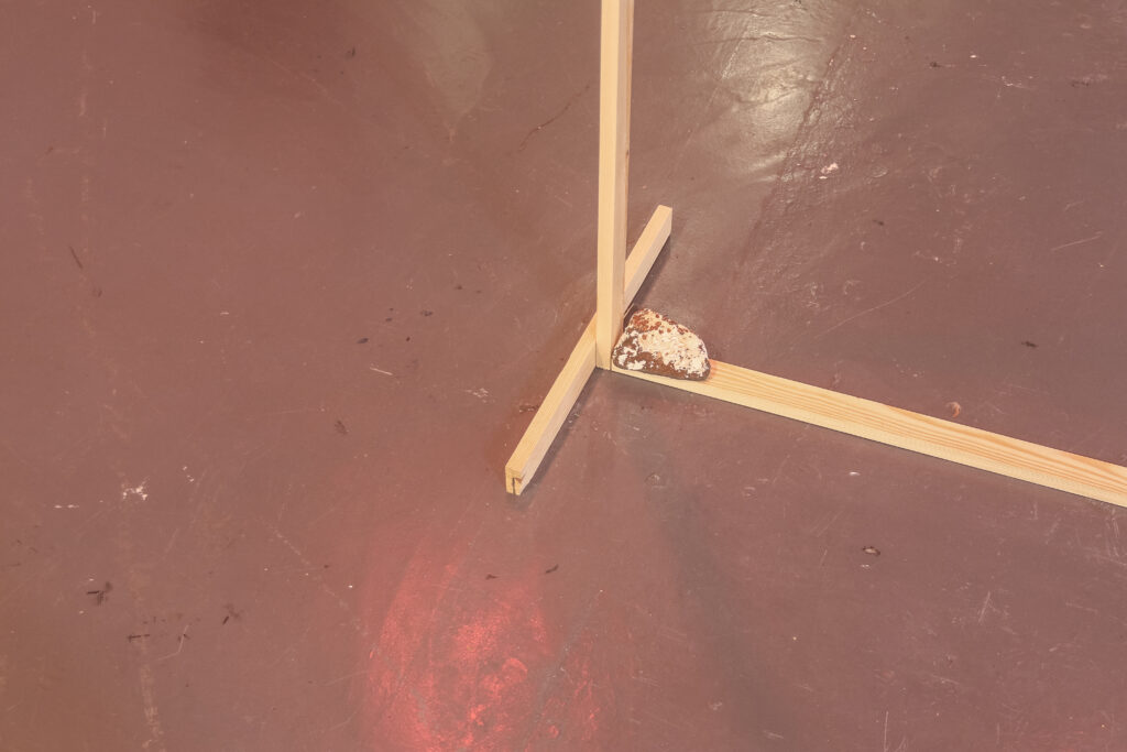 On a grey gallery floor with red lighting is the foot of a thin wooden frame with a rough grey rock with white discolouration on it.