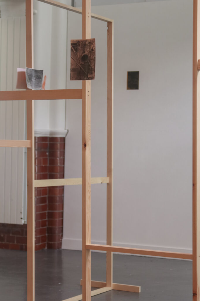 Installation shot of a thin wooden fame with a off-colour hand printed photograph attached to the frame in a white gallery with subtle red lighting and some patches of red brick wall. On the back wall hangs a two photographs next to each other. On the back wall hangs a photograph, and in the foreground on the wooden frame is a photograph of a pipe emerging from a rough wall.