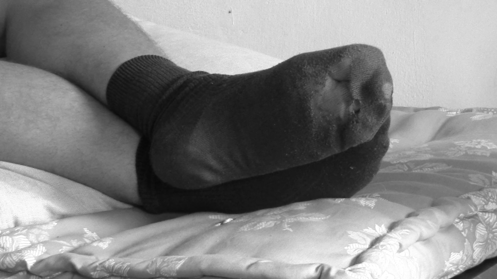 Black and white still of a pair of feet in worn socks on a patterned daybed.