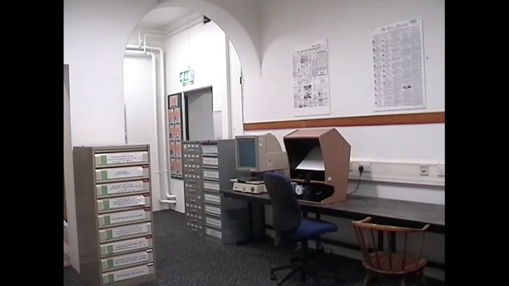 Still of mini DV footage of a room in a library with two microfilm viewers on a grey workbench with a blue office chair and a wooden chair. To the side and adjacent are grey metal filing cabinets.