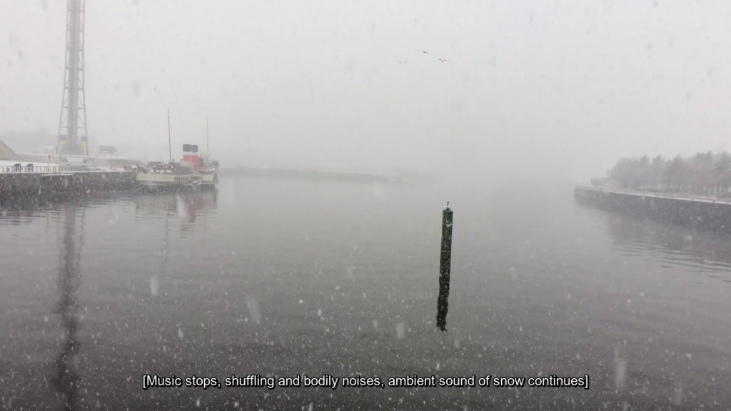 Still of a harbour with heavy snow and limited visibility, at the bottom is the caption [Music stops, shuffling and bodily noises, ambient sound of snow continues].