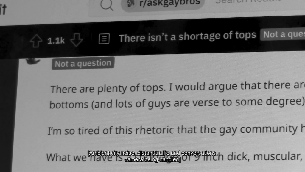 Black and white still of a close up of a computer screen of the webpage r/askgaybros. The topic of the page is ‘There isn’t a shortage of tops’, we can read the lines ‘There are plenty of tops. I would argue that there ar… bottoms (and lots of guys are verse to some degree)… I’m so tired of this rhetoric that the gay community h… What we have is a shortage of 9 inch dick, muscular,’. At the bottom is the caption [Ambient city noise, distant traffic and conversations, camera being handled].