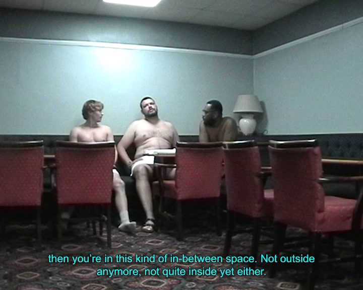 Mini DV footage of three shirtless men of varying skin tones with white towels around their waists, sitting in the corner of a blue pub-style room with red seating and an intricate patterned carpet. Caption reads ‘then you’re in this kind of in-between space. Not outside anymore, not quite inside yet either.’