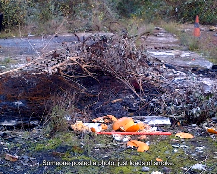 Low-quality BlackBerry phone footage of a bonfire pile made from branches, mattress springs, and debris with a smashed pumpkin in an urban wasteland. Caption reads ‘Someone posted a photo, just loads of smoke’.