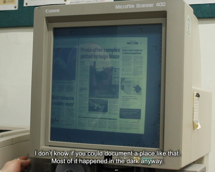 A Canon Microfilm Scanner 400 displaying a newspaper page with the headline ‘Probe after complex gutted by huge blaze’. There is a faint reflection of a face in the screen. The caption reads ‘I don’t know if you could document a place like that. Most of it happened in the dark anyway.’