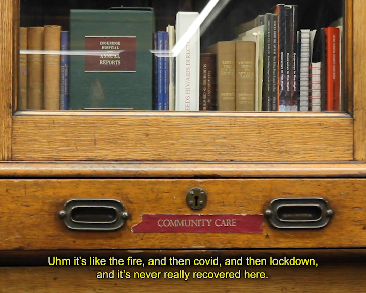 Aged wooden cabinet with a bookshelf behind a glass window door. On a drawer with a keyhole is red tape reading ‘COMMUNITY CARE’. In the cabinet is a range of books; in the middle is a white book spine reading ‘LEEDS HIV/AIDS DIRECTORY’. Caption reads ‘Uhm it’s like the fire, and then covid, and then lockdown, and it’s never really recovered here.’