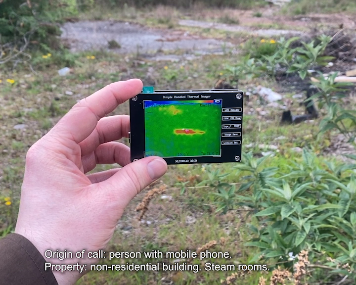 iPhone footage of a light-skinned hand holding a simple handheld thermal imaging device displaying a heat signature in a grassy outdoor area. Caption reads ‘Witness 1: Origin of call: person with mobile phone. Property: non-residential building. Steam rooms.’